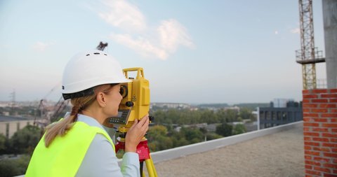 Caucasian beautiful woman topographer in casque measuring angle with total station on roof of building. Female builder. Constructor doing topographic measures. Geodesy concept. Constructing work.