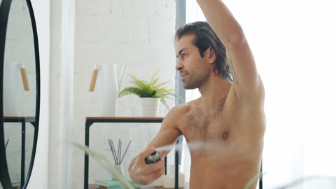 Slow motion of good-looking young man applying deodorant on armpits skin looking at mirror in bathroom during morning routine. People and lifestyle concept.