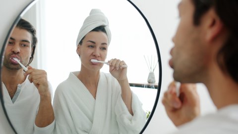 Slow motion of young cheerful man and woman brushing teeth having fun and laughing looking at mirror in bathroom. Relationship and health concept.