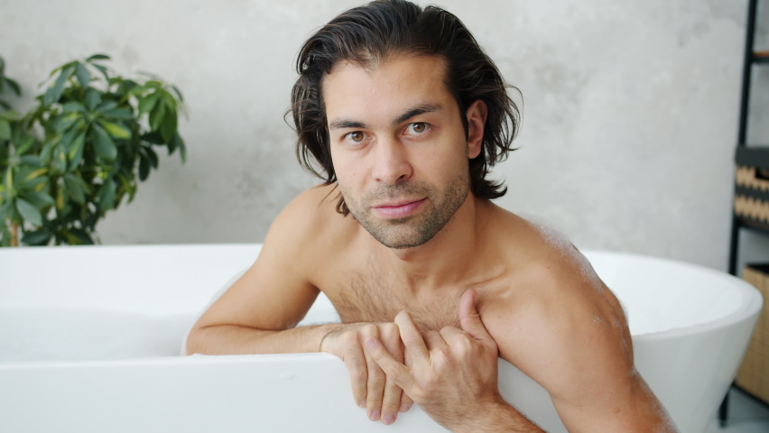 Slow motion portrait of serious young man with stylish long hair looking at camera in bathtub alone at home. Hygiene and spa relaxation concept. Royalty-Free Stock Footage #1061560633