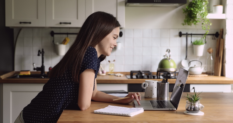Side view pleasant young woman leaning on table, web surfing information on computer, handwriting recipes in kitchen. Smiling 20s female student studying remotely on educational courses at home. | Shutterstock HD Video #1061568655