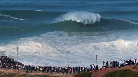 Nazare, Portugal - October 29, 2020: People watching surfers riding giant waves near Praia do Norte beach in Nazare, Portugal. Nazare is famously known for having the largest waves in the world.