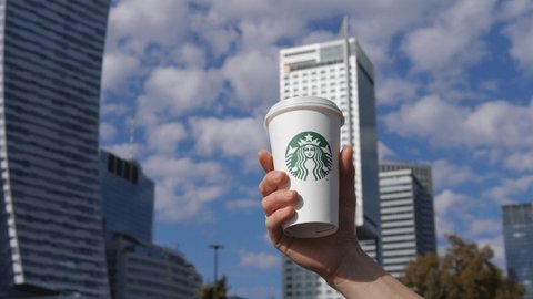 Close up of white hand with Starbucks coffee cup against the background of modern buildings and blue skies. Cafe chain in big city. Warsaw-Poland-2020.