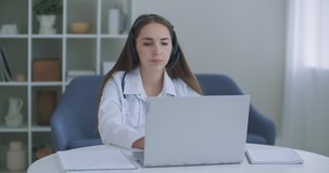 Female medical assistant wears white coat, headset video calling distant patient on laptop. Doctor talking to client using virtual chat computer app. Telemedicine, remote healthcare services concept.
