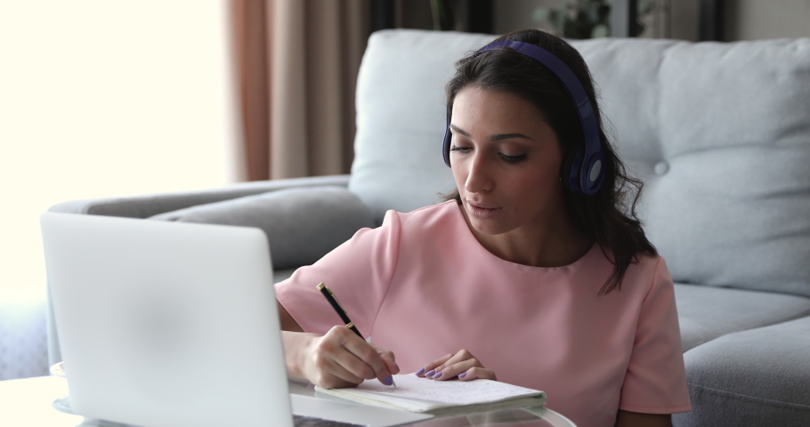 Focused young mixed race indian arabian left-handed woman wearing headphones, listening educational lecture on laptop, writing notes, sitting at coffee table at home, distant e-learning concept. Royalty-Free Stock Footage #1061572714