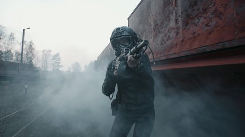 Female surviving future soldier aims with an assault rifle and moves towards the target by rusty train carriage. Lonely cyborg woman in futuristic combat suit goes through cloud of smoke during battle