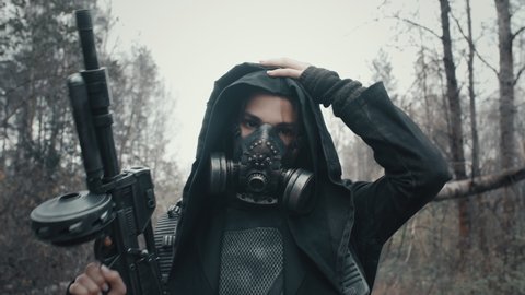 Close-up portrait of woman in grunge clothes hooded mantle and gas mask standing in forest and looking into the camera. Dystopian concept, female future survivor. Post apocalyptic world. Cyberpunk
