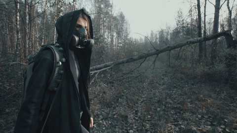 Woman in futuristic military clothes and masks walking along path in dead forest. Stalker concept, female survivor with gun during nuclear or chemical war. Post apocalyptic world. Cyberpunk steam