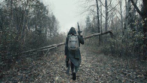 Following shot of a woman in military clothes walking along trail in dark forest. Stalker concept, female survivor with gun during nuclear or chemical war. Post apocalyptic world.  