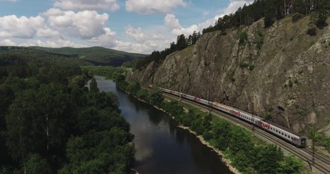 Passenger train an electric locomotive under the rock near river by two-sided winding Trans Siberian railway in the Ural Mountains - Aerial Photography, drone wide into the distance view