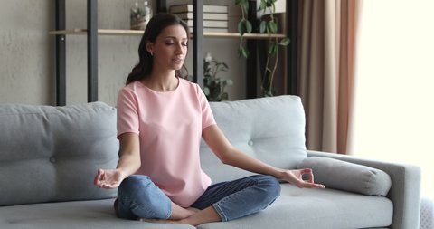 Young calm indian arabian multiracial woman relaxing in lotus position with folded in mudra gesture hands and closed eyes on comfortable sofa, doing yoga breathing exercises, enjoying stress free time