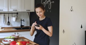 Smiling young pretty woman holding smartphone in hands, searching tasty recipes in mobile cooking application, looking for culinary healthy lifestyle ideas, preparing food alone at home on weekend.