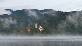 Timelapse video with view of lake Bled and Bled island with Pilgrimage Church of the Assumption of Maria under fog and mist rises above the water in early morning, Bled, Slovenia