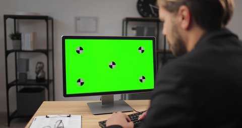 View from shoulder of bearded man in formal wear sitting at table and working on computer with chroma key screen. Concept of business and technology.