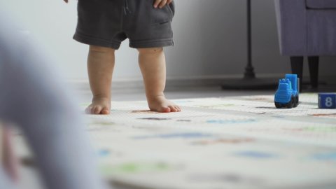 Little boy child, gray shorts white T-shirt stands home hall children room soft safe carpet among toys. First steps, confident gait small bare feet. Lesson nanny, educator, parents. Develop child home