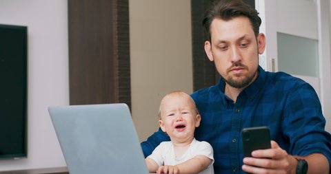 Young bearded man sitting at desk at home with laptop, holding cute baby at knees, writing on keyboard, reading message on smartphone. Baby makes grimaces and shows dissatisfaction. Slow motion