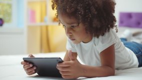 Adorable afro kid girl using digital tablet watching cartoons lying on bed at home. Close up portrait of cute african child playing game on tablet pc relaxing in bedroom - Powered by Shutterstock - Get 15% off with code: PIKWIZARD15