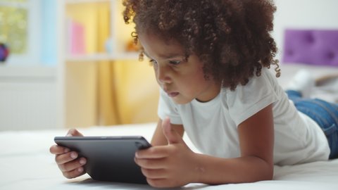 Adorable afro kid girl using digital tablet watching cartoons lying on bed at home. Close up portrait of cute african child playing game on tablet pc relaxing in bedroom
