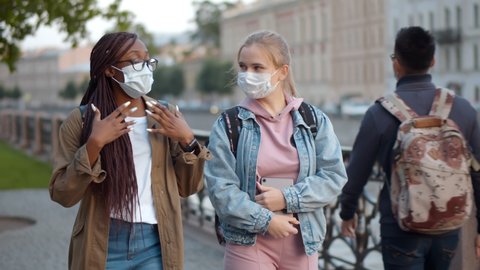 Multinational student women in medical mask talking while walking down city street. African and caucasian young female in safety mask strolling in city and chatting