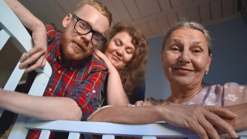 Bottom view of mother, father and grandmother watching at sleeping baby in cot. Happy smiling young couple and elderly woman looking at newborn child in crib