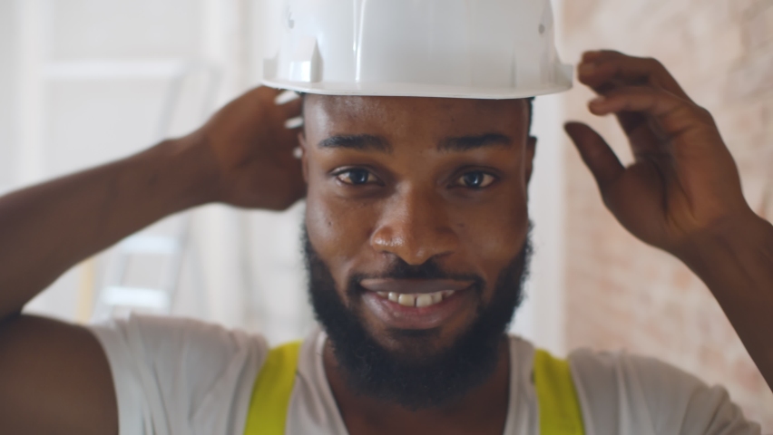 Close up portrait of african manual worker putting on safety helmet and smiling at camera. Afro-american migrant worker in uniform doing house renovation | Shutterstock HD Video #1061585554