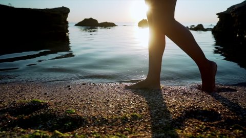 Legs of young girl walking barefoot on wet sand island beach, sun lens flare. Beautiful feet of slim woman near sea on sunset or sunrise. Slow motion.