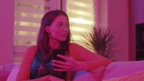 Fabulous young Caucasian woman looking around and liking photos of guys in dating up. Secrets concept. Alone in living room. Attractive girl using smartphone, touching screen. Pink neon light. Home