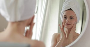 Attractive woman rapped in towel after shower applying moisturizing cream on her face while looking at mirror. Concept beauty routine and health care.