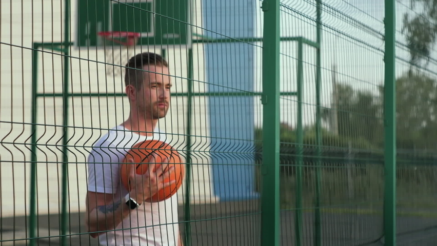 Thoughtful basketball player with a ball looking through the metal fence of a court. He feels the lack of motivation to continue