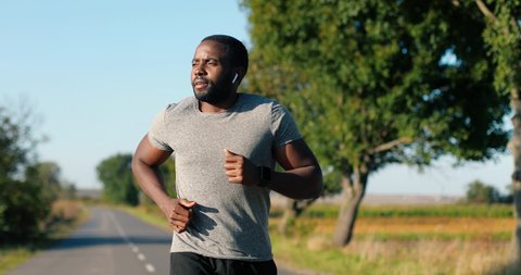African American handsome strong male jogger in headphones running on road in countryside on summer day. Attractive sporty man jogging outdoors. Sport concept. Sportsman runner. Morning nature.