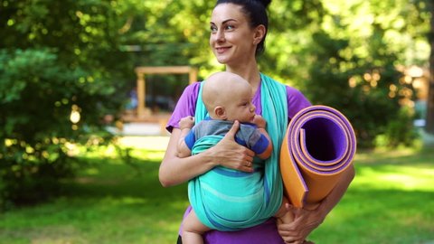 Smiling woman with hair bun walking in park and carrying baby in sling, soft mat under her arm. Tracking shot fit mom going to yoga class with little child. Concept of sport