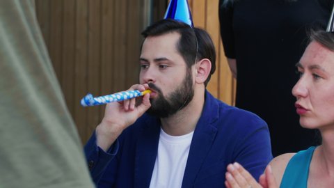 Close-up young Caucasian man tired of celebrating birthday party with friends, wearing funny hat and blowing whistle.