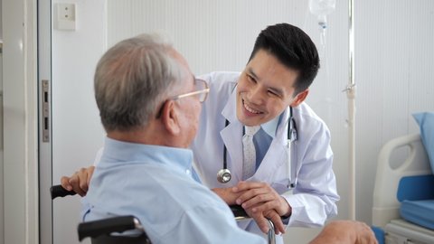 Happy Asian male doctor communicating with elder patient in wheelchair at clinic. Elderly healthcare concept. Slow motion
