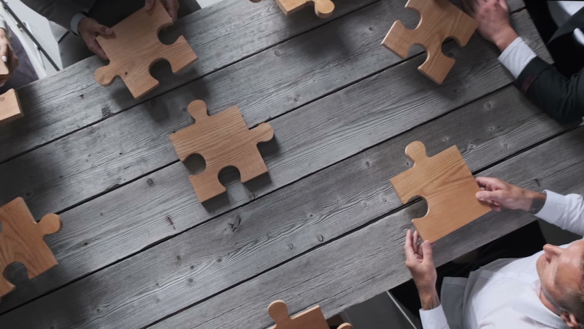 Business people team sitting around meeting table and assembling wooden jigsaw puzzle pieces unity cooperation ideas concept | Shutterstock HD Video #1061596747