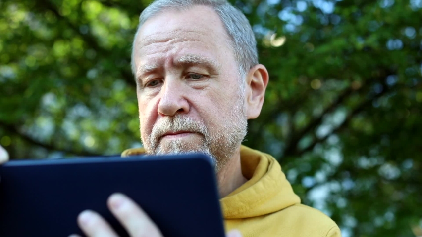 Senior bearded man using tablet for communication looking at screen resting outdoor enjoying autumn spring sunny nature. Sunny background. Internet connect. Free communicating with family.