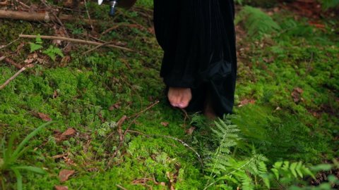 Bare female's feet. Legs of a woman in long black dress walking on green soft grass. Close-up. Slow motion.