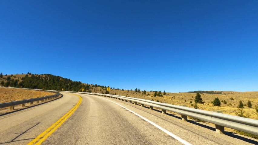 Driving Highway 287 Curving weathered two-lane asphalt highway in southwest Montana passing dry amber autumn grass and green pine trees under a clear blue sky-Drive Plate-POV