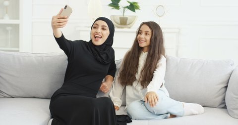 Beautiful young Arabian mother in hijab sitting on couch wih cute little daughter and taking selfie photos with smartphone camera. At home. Woman with kid making pictures selfies with mobile phone.