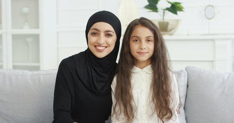 Portrait of beautiful young muslim mother in headscarf smiling to camera while sitting with cute teen daughter on sofa in living room. Pretty Arabian woman in hijab and small teenage girl on couch.
