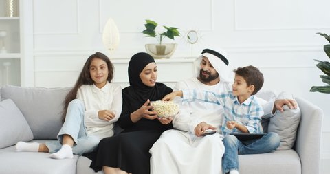 Cheerful Arabian parents with little children sitting on couch, smiling, watching movie on TV and eating popcorn. Cute Arab small kids with mother and father spending time together at home.