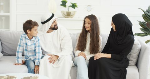 Portrait of cheeful Muslim family with two kids sitting on couch and smiling joyfully to camera. Small cute boy and girl with mother and father at home. Arabian parents with little children hugging.