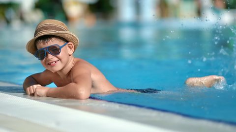 Trendy male kid splashing in swimming pool having fun and positive emotion slow motion. Smiling playful little boy in straw hat and sunglasses enjoying summer travel vacation. 4k Dragon RED camera