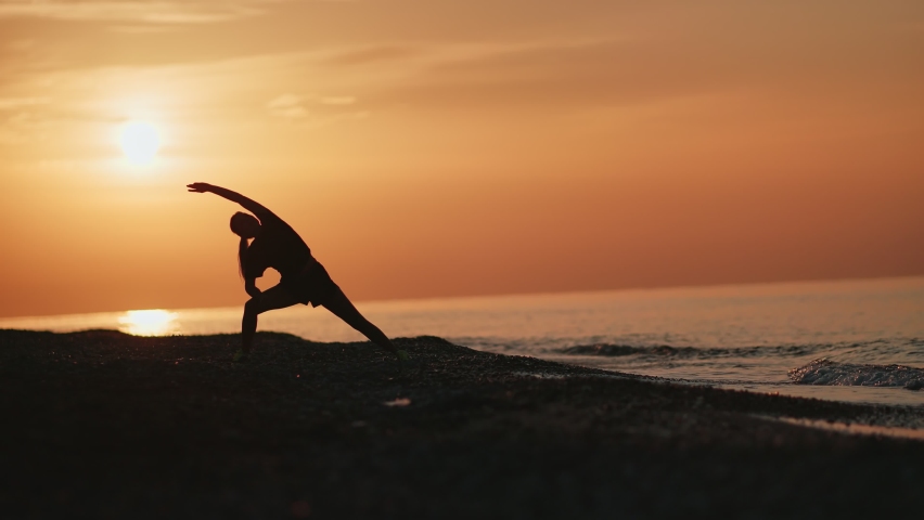 Silhouette of fitness woman practicing asana yoga pose on beach at dramatic sunrise sky. Female doing sports exercise stretching body at seascape morning nature long shot. 4k Dragon RED camera Royalty-Free Stock Footage #1061606293