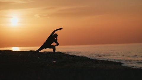 Silhouette of fitness woman practicing asana yoga pose on beach at dramatic sunrise sky. Female doing sports exercise stretching body at seascape morning nature long shot. 4k Dragon RED camera