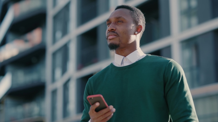 Confident african american man using mobile phone in modern city district. Positive business guy browsing financial news smiling standing outdoor. Successful adult. | Shutterstock HD Video #1061608948