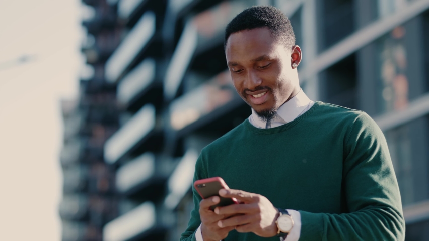 Confident african american man using mobile phone in modern city district. Positive business guy browsing financial news smiling standing outdoor. Successful adult. | Shutterstock HD Video #1061608948