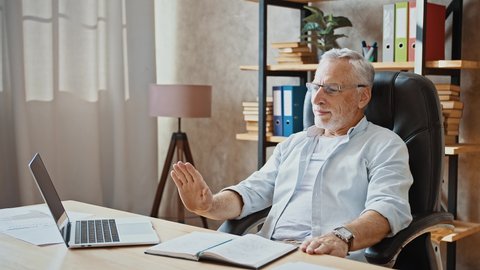 Mature businessman in glasses is putting hands behind his head and smiling being happy while sitting at table with laptop and notepad on it. Working at home office. Remote job, freelance. Slow motion