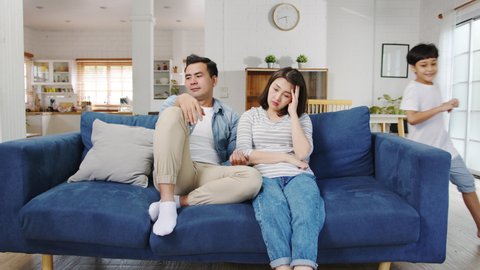 Asia family dad and mom sit on couch feels annoyed exhausted while daughter and son have fun shouting run around sofa in living room at home. Social distancing, Quarantine for corona virus prevention.