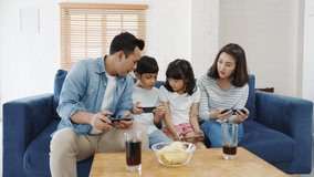 Happy Asian family dad, mom and kids playing funny game online on smartphone sit sofa in living room at house. Self-isolation, stay at home, social distancing, quarantine for coronavirus prevention.