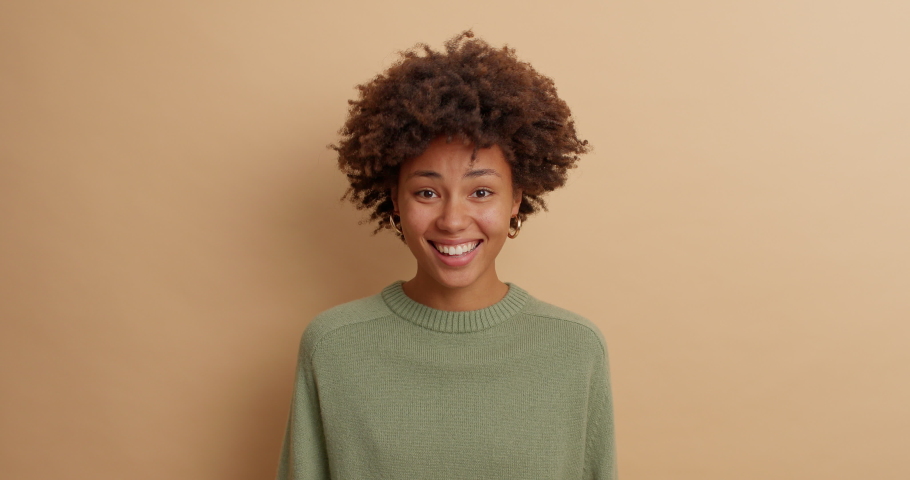 Curious dark skinned woman asks what and nods head with agreement says thats true agrees with you smiles broadly shows white perfect teeth dressed in casual jumper isolated over beige background | Shutterstock HD Video #1061613004